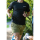 En person jogger på en sti i LiiteGuards Ground-Tech T-shirt, Herre, Black og olivenfarvede shorts. Den svedtransporterende tee holder dem komfortable midt i det grønne, hvor kun deres overkrop og ben er synlige.