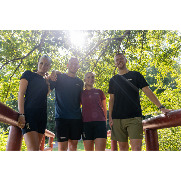Fire personer står side om side på en bro i det fri og smiler til kameraet. Sollyset filtreres gennem grønne blade, og de er iført afslappet sportstøj, herunder LiiteGuard Ground-Tech T-shirt til mænd i sort.