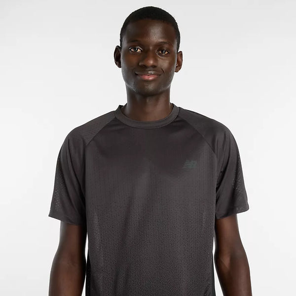 En mand står på en almindelig lysegrå baggrund iført en sort New Balance Viz Tech Ss kortærmet T-shirt til mænd, designet til træning. Han ser fremad med et neutralt udtryk.