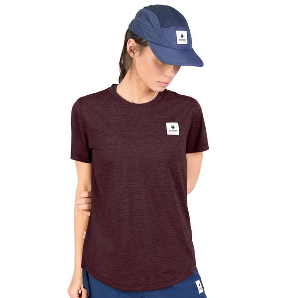 En person iført en Saysky Clean Combat Pro-T-shirt, Dame, i farven burgundy med et logo-mærke, kombineret med blå shorts og en blå kasket med et matchende logo-mærke, står med den ene arm bag ryggen og kigger ned.