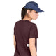 En kvinde står med ryggen til kameraet iført en Saysky Clean Combat Pro T-shirt i farven Dame, Burgundy og en marineblå kasket. Hun har sit lange, mørke hår sat op i en hestehale og kigger over skulderen. Den hurtigtørrende T-shirt sikrer komfort, mens den hvide baggrund fremhæver hendes sporty stil.