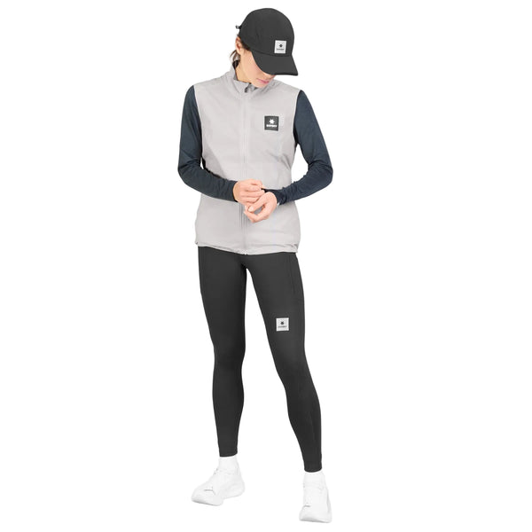 En person iført en sort kasket, en grå Saysky W Clean Pace Flex Vest til kvinder, en sort langærmet trøje med paneler af stretchmesh, sorte leggings og hvide sneakers står foran en hvid baggrund og ser ned med hænderne foldet foran sig.