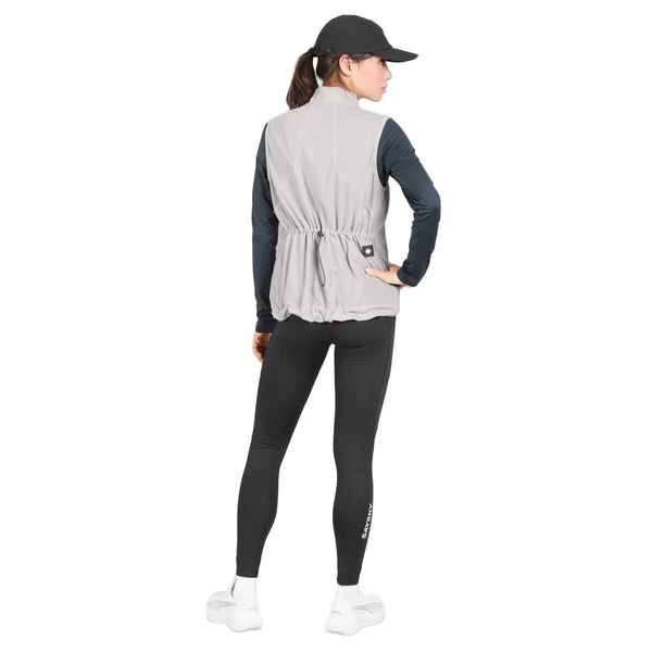 En kvinde iført Saysky W Clean Pace Flex Vest, Dame, grå – med elastiske mesh-paneler, der sikrer god åndbarhed – samt en sort langærmet bluse, sorte leggings, hvide sneakers og en sort kasket står med ryggen til og den ene hånd på hoften.