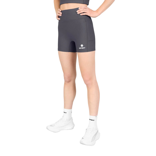 En person står med hænderne i siden og har Saysky W Combat+ Rib Short Tights 4″ til kvinder i gråt på – fremstillet af åndbart ribstof – kombineret med en matchende top, hvide sokker med sort tekst og hvide sportssko på en ensfarvet hvid baggrund.