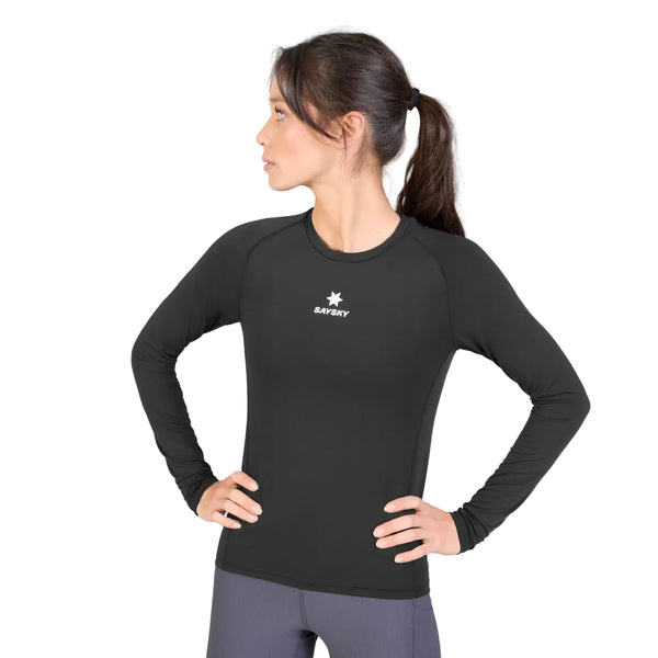 En kvinde med mørkt hår i hestehale har en Saysky W Combat Tech Long Sleeve, Dame, Black på – en sort langærmet sportstrøje, der er kombineret med grå leggings. Hun står med hænderne på hofterne og kigger til venstre. T-shirten er fremstillet af svedtransporterende løbetøj og har en lille hvid stjerne samt Saysky-logoet på brystet.