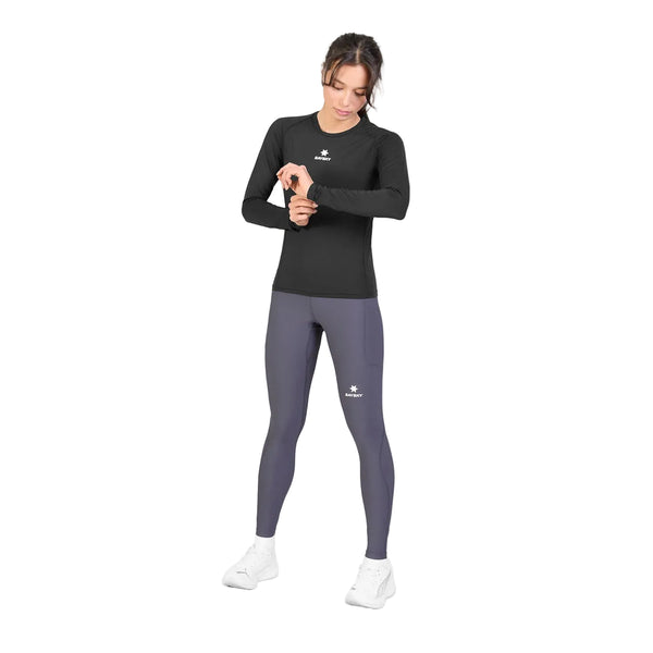 En kvinde står foran en hvid baggrund iført den sorte Saysky W Combat Tech Long Sleeve til kvinder, kombineret med grå leggings og hvide sko. Hun kigger på sit håndled, som om hun tjekker sit ur. Dette svedtransporterende løbetøj fra Saysky sikrer, at hun forbliver komfortabel og tør under enhver aktivitet.