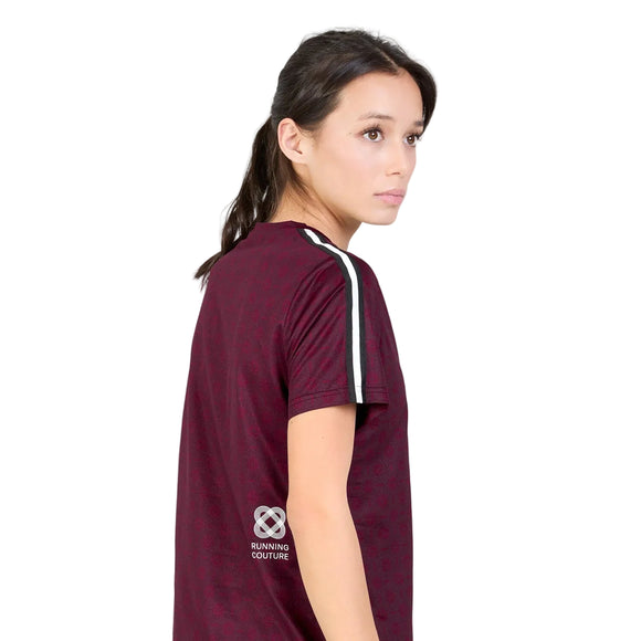 En kvinde med mørkt hår i en hestehale har en Saysky W Couture Combat T-shirt på, Dame, Burgundy Aop, som har hvide striber på ærmerne og teksten »Running Couture« med et logo trykt tæt på nederkanten. Hun kigger til højre foran en hvid baggrund.