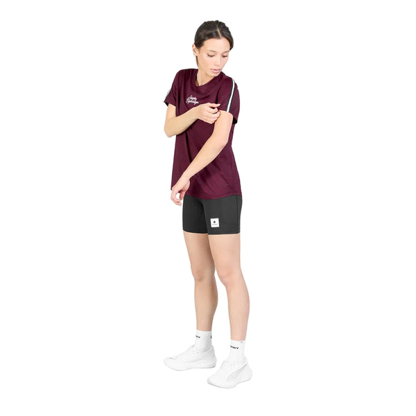 En person står på en hvid baggrund iført en Saysky W Couture Combat T-shirt, Dame, Burgundy Aop, kombineret med sorte shorts, hvide sokker og hvide sko. Personen kigger ned og rører ved sin overarm med den modsatte hånd.