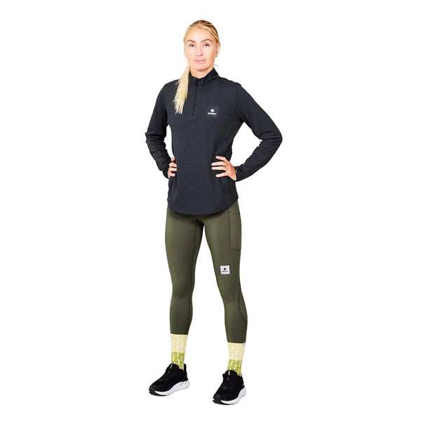 En person står foran en ensfarvet lysegrå baggrund iført Saysky W Blaze Half Zip Light-Weight Fleece til kvinder i sort – fremstillet af letvægts-løbefleece af genanvendt polyester – kombineret med olivengrønne leggings med lommer, gule sokker og sorte løbesko. Med hænderne på hofterne og blikket rettet fremad udstråler vedkommende et selvsikkert udtryk, der passer perfekt til aktive kvinder.