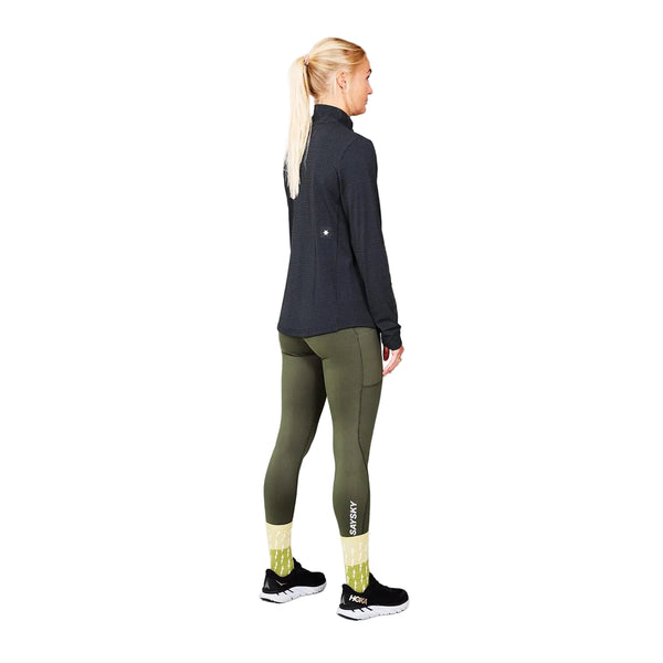 En kvinde med blondt hår i hestehale står med ryggen til; hun er iført en mørk langærmet bluse af genbrugt polyester, olivengrønne leggings med SAYSKY-logo, gule mønstrede strømper og sorte løbesko. Hendes stilfulde outfit til kvinder fuldendes af den funktionelle Saysky W Blaze Half Zip Light-Weight Fleece, Dame, Black – en elegant, letvægts løbefleece, der fremstår tydeligt mod en ensfarvet grå baggrund.