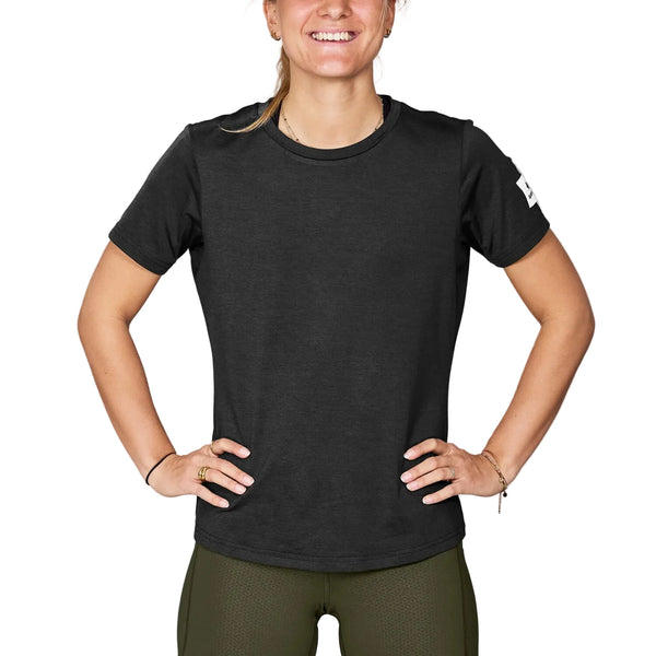 En person står med hænderne i siden, smiler og har en Saysky W Clean Pace T-shirt, Dame, sort, på, kombineret med olivengrønne bukser. Ansigtet er delvist synligt på en ensfarvet hvid baggrund.