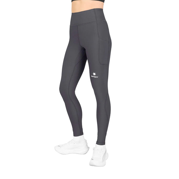 Man ser en person iført et par højtaljede W Combat+ Rib Long Tights, Dame, grå fra Saysky, med sidelommer, fremstillet af behageligt ribstof. Strømpebukserne har et lille hvidt stjerne-logo og ordet »TERRY« på det øverste venstre lår. Kun underkroppen og benene er synlige. Outfitet er kombineret med hvide sneakers.