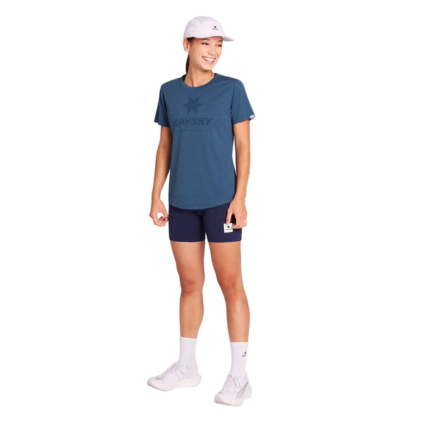 En person står foran en hvid baggrund iført en blå Saysky W Logo Combat Pro-T-shirt til kvinder, marineblå shorts, hvide sokker, hvide sko og en hvid kasket. Personen smiler og holder et lille kort i venstre hånd og ser afslappet og stilfuld ud i sit hurtigtørrende træningstøj.
