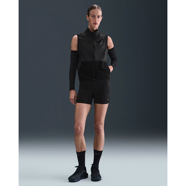 En person står med front mod en almindelig mørk baggrund iført en sort vest uden ærmer med lynlås, Nike Dri-FIT High-Waisted 4" Tight Running Shorts til kvinder i sort, sorte sokker, sorte sko og lange sorte ærmer.
