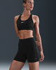 En kvinde står på en mørk baggrund iført en sort Nike sports-bh og Dri-FIT High-Waisted 4" Tight Running Shorts for women i Black/Black, mens hun kigger lidt til højre med venstre hånd hvilende på låret.