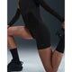 En person har Nike Dri-FIT High-Waisted 4" Tight Running Shorts, Dame, Black/Black på, mens han knæler på det ene knæ og strækker det andet ben ved at holde foden bag sig mod en almindelig mørk baggrund.