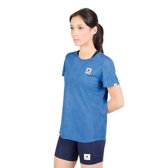 En person med langt, mørkt hår, iført en Saysky W Reactive Flow T-shirt, Dame, Blue Aop og mørkeblå shorts, står foran en hvid baggrund med hænderne bag ryggen.
