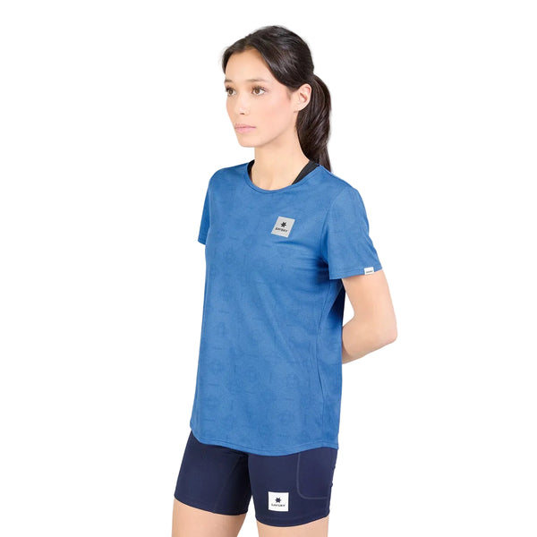 En person med langt, mørkt hår, iført en Saysky W Reactive Flow T-shirt, Dame, Blue Aop og mørkeblå shorts, står foran en hvid baggrund med hænderne bag ryggen.