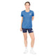 En person står på en hvid baggrund iført en Saysky W Reactive Flow T-shirt, Dame, Blue Aop, sammen med marineblå shorts, hvide sokker og hvide sko. Personen smiler, med den ene arm bag ryggen og den anden afslappet ned langs siden.