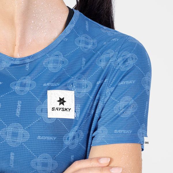 En person iført Saysky W Reactive Flow T-shirt, Dame, Blue Aop – en blå, kortærmet, svedtransporterende T-shirt med et geometrisk mønster og et hvidt Saysky-logo på brystet. Deres hud er dækket af vanddråber.