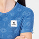 En person iført Saysky W Reactive Flow T-shirt, Dame, Blue Aop – en blå, kortærmet, svedtransporterende T-shirt med et geometrisk mønster og et hvidt Saysky-logo på brystet. Deres hud er dækket af vanddråber.