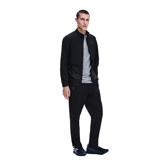 En person står med ansigtet vendt fremad og er iført en sort sportsjakke, et par On Weather Pants, Herre, Sort, en grå T-shirt og sorte sneakers. De vandafvisende bukser og jakken er perfekte til at trodse vejret under udendørs træning. Baggrunden er ensfarvet hvid.
