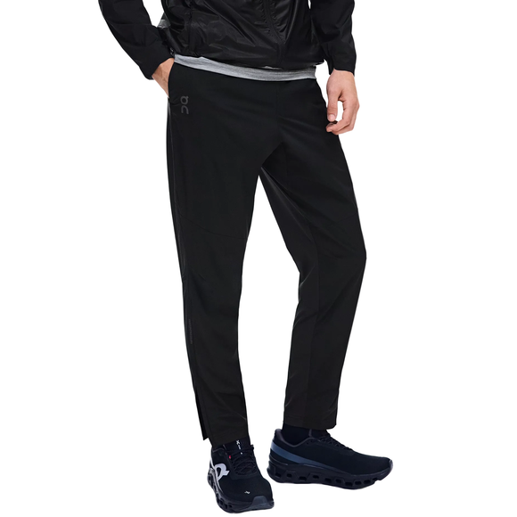 En person har On Weather Pants, Herre, Black på, sorte sneakers med hvide såler, en grå skjorte og en sort jakke. Personen står med den ene hånd i lommen foran en hvid baggrund.
