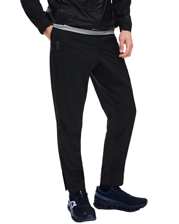 En person har On Weather Pants, Herre, Black på, sorte sneakers med hvide såler, en grå skjorte og en sort jakke. Personen står med den ene hånd i lommen foran en hvid baggrund.