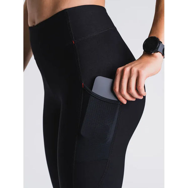 En kvinde i Fusion 3/4 Training Tights, Dame, SuperBlack stikker sin smartphone ned i netlommen på siden. De svedtransporterende tights matcher et sort armbåndsur, alt sammen på en lys baggrund.