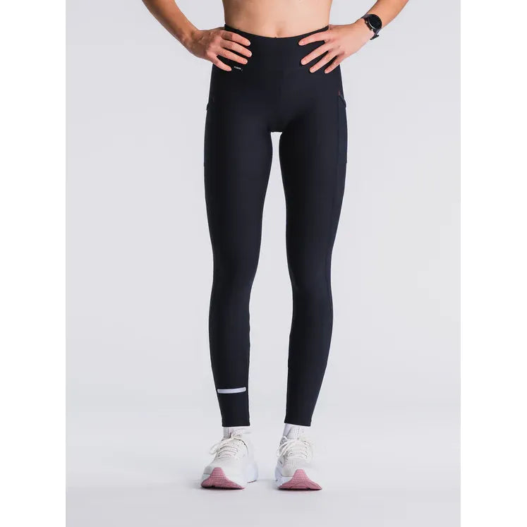 En kvinde med hænderne på hofterne bærer Fusions SuperBlack Dame Training Tight-leggings, et smartwatch og hvide sneakers. Billedet er beskåret ved brystet og placeret på en almindelig hvid baggrund.