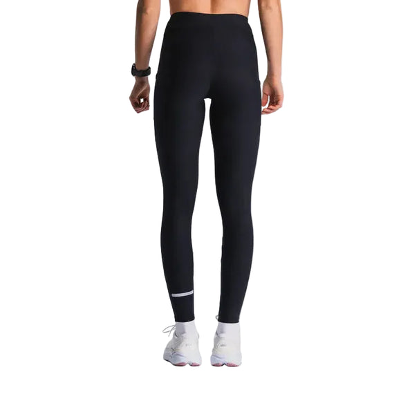 En person står med front mod kameraet iført Fusion Training Tight, Dame, SuperBlack-leggings, et sort armbånd og hvide sportssko på en almindelig lys baggrund.