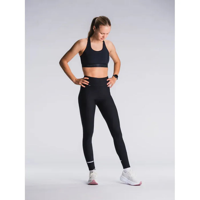 En kvinde står på en almindelig lys baggrund iført Fusion's Training Tight Dame SuperBlack højtaljede leggings, en sort sports-bh og hvide sportssko. Hun kigger ned med hænderne på hofterne.