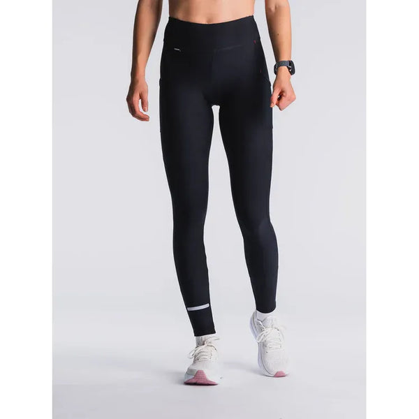 En kvinde iført Fusion Hot X-Long Tights, Dame, Black med en lille refleksstribe på venstre ben, et armbåndsur og hvide løbesko står på en almindelig lys baggrund.