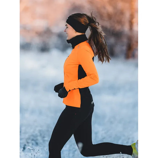 En kvinde løber udendørs i sneen i Fusion Hot X-Long Tights, Dame, Black, sammen med en orange jakke, handsker og et sort pandebånd. Den snedækkede baggrund er sløret med synlige træer.