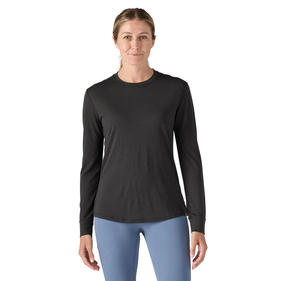 En kvinde står og kigger fremad iført en sort Patagonia L/S Cap Cool Merino Blend Graphic Shirt, Dame og lyseblå bukser på en hvid baggrund.