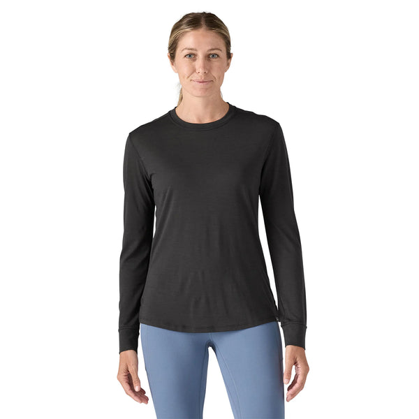 En kvinde står og kigger fremad iført en sort Patagonia L/S Cap Cool Merino Blend Graphic Shirt, Dame og lyseblå bukser på en hvid baggrund.