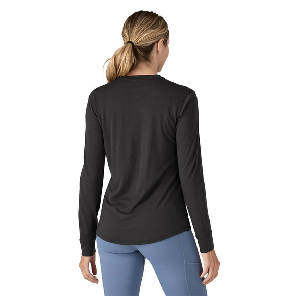 En kvinde med lysebrunt hår i hestehale er iført en sort Patagonia L/S Cap Cool Merino Blend Graphic Shirt, Dame og lyseblå bukser. Hun kigger væk fra kameraet mod en hvid baggrund.