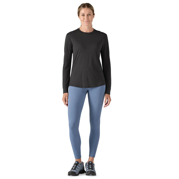En kvinde står med front mod en hvid baggrund iført en Patagonia L/S Cap Cool Merino Blend Graphic Shirt i sort, lyseblå leggings og sorte sportssko.