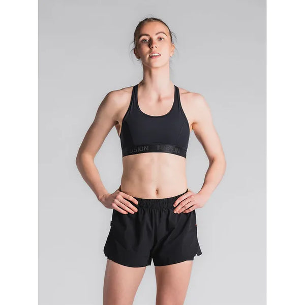 En kvinde poserer på en almindelig lysegrå baggrund iført Fusion Run Shorts, Dame, Black og en matchende sports-bh, med hænderne på hofterne og et neutralt udtryk, klar til sin næste løbetur.