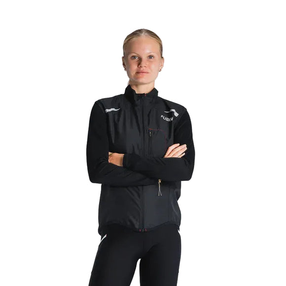 En kvinde med tilbagestrøget blondt hår står på en almindelig baggrund iført Fusion S1 Run Vest, Dame, Black, og sorte bukser med korslagte arme og kigger ind i kameraet.