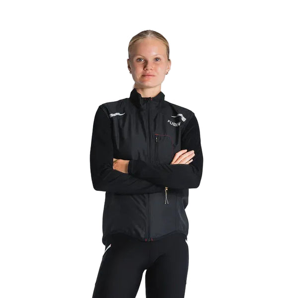 En kvinde med tilbagestrøget blondt hår står på en almindelig baggrund iført Fusion S1 Run Vest, Dame, Black, og sorte bukser med korslagte arme og kigger ind i kameraet.