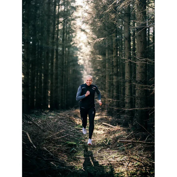 Iført Fusion S1 Run Vest til kvinder i sort bevæger en løber sig langs en smal skovsti omgivet af høje træer og sollys, der filtrerer gennem grenene.