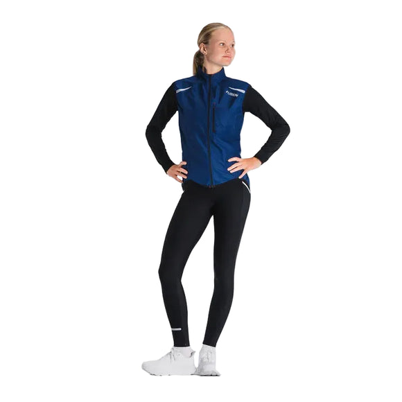 En person står på en lys baggrund iført Fusion S1 Run Vest, Dame, Night Blue over en sort langærmet top, sorte leggings med hvide detaljer og hvide sportssko med hænderne på hofterne.