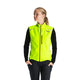 En person står let vendt og er iført Fusion S1 Run Vest til kvinder i gul over en sort langærmet skjorte og bukser på en almindelig lysegrå baggrund.