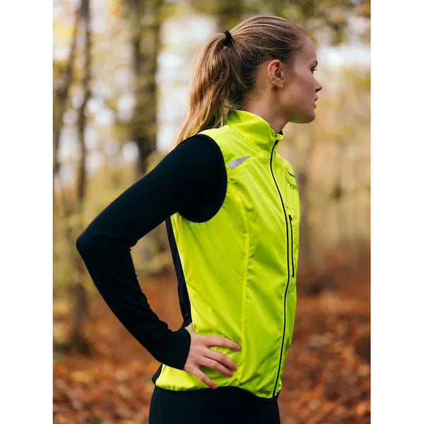 En person med hestehale står udendørs iført Fusion S1 Run Vest, Dame, Yellow over en sort langærmet skjorte. Hænderne hviler på hofterne, mens de kigger til siden med efterårstræer i baggrunden.