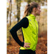 En person med hestehale står udendørs iført Fusion S1 Run Vest, Dame, Yellow over en sort langærmet skjorte. Hænderne hviler på hofterne, mens de kigger til siden med efterårstræer i baggrunden.