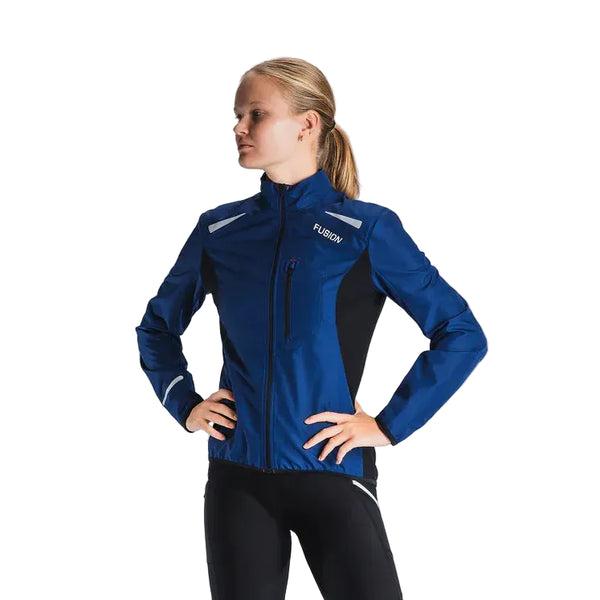 En person iført Fusion S1 Run Jacket til kvinder i Night Blue står med hænderne på hofterne og kigger til siden mod en almindelig lys baggrund.
