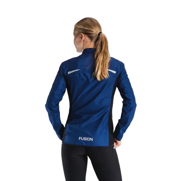 En kvinde med langt blondt hår i hestehale bærer Fusion S1 Run Jacket til kvinder i Night Blue med "FUSION" trykt på den nederste del af ryggen. Hun står med ryggen til kameraet på en almindelig baggrund.