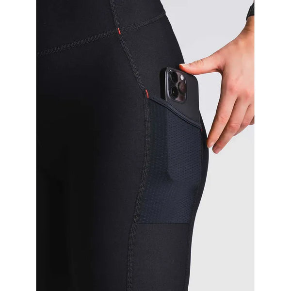 Nærbillede af en person iført Fusion Short Training Tights, Dame, SuperBlack, der lægger en smartphone i netlommen på låret mod en ensfarvet baggrund.