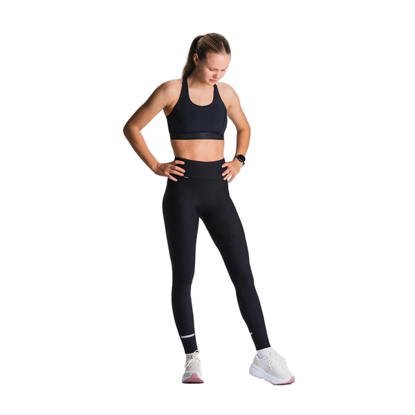 En kvinde står med hænderne på hofterne og kigger ned iført en sort sports-bh, Fusion X-Long Training Tight Dame i SuperBlack (svedtransporterende) og hvide sportssko. Hendes hår er sat op i en hestehale. Baggrunden er hvid.